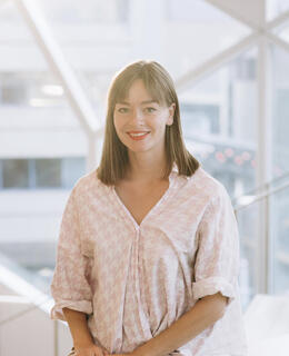 In a well-lit room stands a young white woman with dark blonde hair reaching her shoulders. She smiles, wearing a light purple shirt and jeans. The background is a brightly lit room with soft lighting.