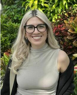 Isabel, wearing black frame glasses, beige sleeveless mock neck shirt and black cardigan, stands in front of green foliage. 
