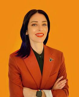 Image of a smiling person with dark hair wearing a rust coloured suit jacket with their arms crossed.
