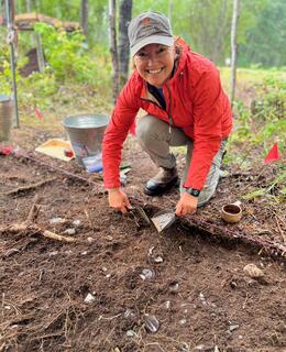 This image shows a person kneeling in the dirt. They are wearing a bright pink jacket, beige cargo pants, and a grey hat with an Athabasca University logo. They are holding a trowel in their right hand and are scraping at the dirt. There are many pieces of pottery and other artifacts scattered around them. In the background, there are trees and a blue sky.