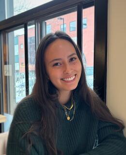 Claire: brown hair, olive skin, brown eyes. Wearing a green sweater sitting at a table in a restaurant, smiling at the camera.  