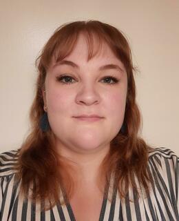 A headshot of a Caucasian female with medium-length brown hair and bangs, wearing a blue and white striped shirt. 
