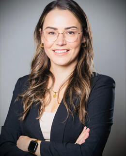 Woman with long brown hair wearing wire framed glasses in a black blazer and beige top smiling with her arms crossed