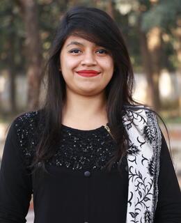 A woman with long dark hair and bright red lipstick is smiling at the camera. She is wearing a black top with a sequined design on the front and a black-and-white patterned scarf draped over her shoulder. The background shows a park with trees and blurred foliage.