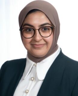 This is a headshot of Dr. Mian Akram who is a woman wearing a hijab, glasses, white shirt, and blazer. She is smiling at the camera.
