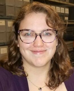 A headshot of a white woman with light brown shoulder length wavy hair and light blue eyes framed by purple cat eyed glasses is smiling at the camera. Behind her are shelves of grey archival boxes.