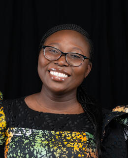 Professional headshot of Motilola against a black background, wearing a tie-dye dress and smiling.