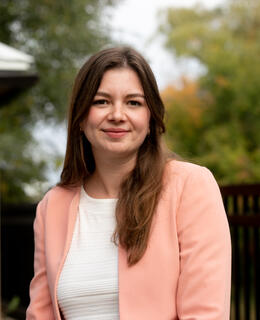 Dr. Chorlton is wearing a white blouse and pink jacket with her hair down. She is smiling in an front of a blurred, outdoor scene.