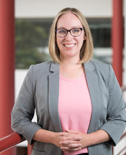 A picture of a woman in a pink shirt and grey blazer, standing in front of two red pillars.
