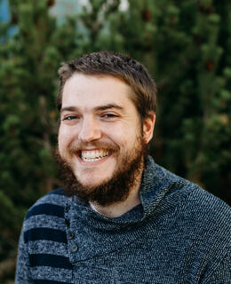 Headshot of a mid 30s white man, with short brown hair and medium length beard smiling outside.