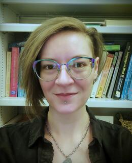 A professional looking head shot of a smiling person with shorter brown hair and multi-coloured glasses in front of a bookshelf filled with Linguistic textbooks.