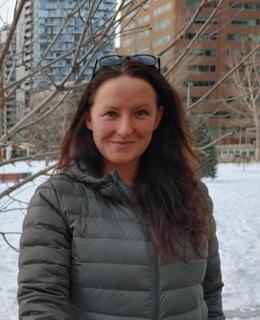 Close-up photo of Jessica, standing outside in the snow with buildings and bare trees in the background. She is wearing a green jacket, has long brown hair and glasses on her head.