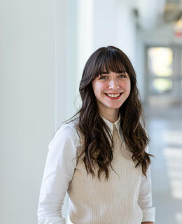 Person standing in a sunny, long, white hallway. Sydney is Caucasian, has long brown hair, and is wearing a big smile.  