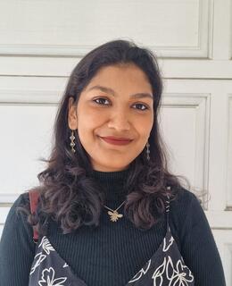 A picture of Anuneeta wearing a black dress and a small bee necklace infront of a white textured background