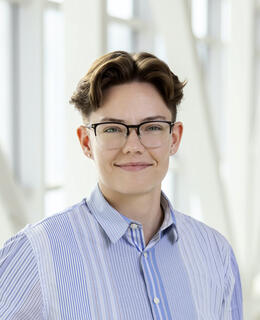 An image of a person with short brown hair and glasses smiling at the camera. They are wearing a blue striped collared shirt.