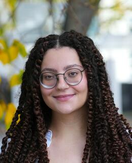 Headshot of Nada Hassanin standing in front of a tree in fall with yellow leaves, wearing glasses, a white button down top, and hair in long brown and rust coloured twists