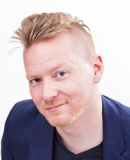 Image of a smiling man with short blonde hair. He is wearing a dark blue jacket over a black t-shirt.