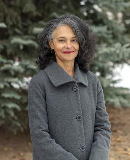 A photo of Dr. Suzette Mayr in grey coat in front of pine trees.