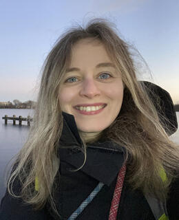 The image shows a person smiling outdoors near a large body of water. The background includes a pier or dock and distant trees. The sky is clear with soft hues, suggesting late evening light.