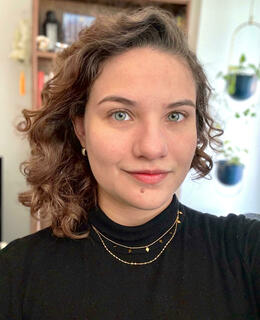 A person with curly hair wearing a black turtleneck, smiling softly, with a bookshelf and plants in the blurred background.