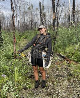 Alice Kennedy photographed in the field. She stands in a forest wearing a black raincoat and planting bags. She rests her hand on her planting shovel and smiles.