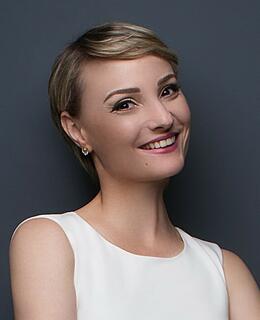 A woman with short, light brown hair, confident smile is the subject of the close-up studio shot. She is positioned slightly angled to her right, facing the camera. Her expression is approachable, friendly, and engaging. She wears a simple, off-white sleeveless dress. The background is a uniform, neutral dark gray, which smoothly fades. The overall impression is of a professional, friendly portrait.