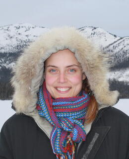 Dr. Allyson Menzies, standing in front of a mountain on a winter landscape, wearing a hooded jacket and a Metis sash