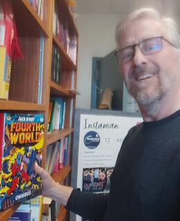 Picture of Dr. Mather standing in front of a bookcase holding a comic titled “Fourth World”. Checking out Kirby