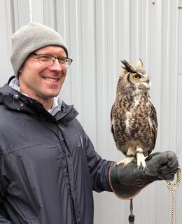 A smiling man with glasses in a grey hat and charcoal windbreaker, with an owl perched on his left arm