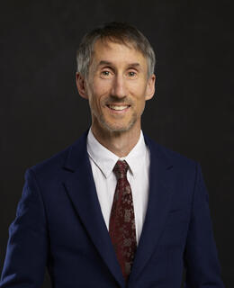 Image of Dr. Michael Tavel Clarke. He has short hair and wears a navy-blue suit and red tie, and stands in front of a dark background.