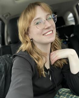 Picture of white woman smiling and sitting in a car. She has medium length light hair and a black shirt, with green plaid pants on. She is wearing round metal glasses.