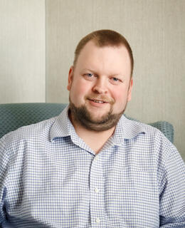 Portrait photo of Jason Reid -- a man wearing a blue and white checked shirt is seated comfortably in a chair, appearing relaxed and contemplative.