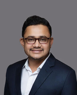 The image appears to be a professional headshot of a man wearing glasses, a dark blazer, and a white shirt, with a neutral gray background. He has a confident and approachable expression.