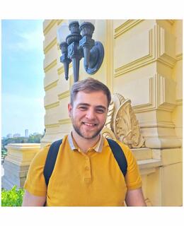 White male smiling with a mustard shirt and a backpack, against the backdrop of a pastel-yellow building and a blue sky.