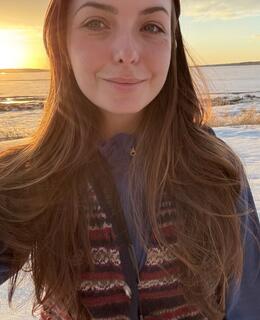 Subject is smiling with a sunset in the background. Subject has long brown hair and is wearing a blue fleece with a short-sleeved wool grandpa sweater layered on top.