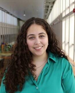 A professional photo of Sandra Amin smiling warmly, with a blurred background in the Taylor Institute building at the University of Calgary.