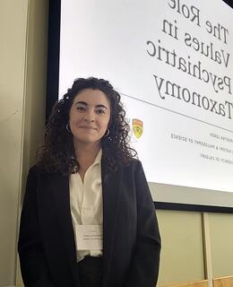 Christina stands in front of a projector screen wearing a blazer and a name tag. She has brown curly hair and brown eyes.