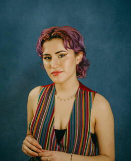 Sofia, a lean person with fair complexion stands in front of a textured blue background. Their hair is curly and short, in a pixie length mullet and dyed purple. They're wearing a striped vest with a rainbow pattern. Their hands are crossed in front of their chest. 