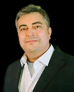 A head-and-shoulders studio portrait of Dr. Javier Alfaro  set against a plain black background. He has short, neatly combed salt-and-pepper hair, thick eyebrows, and a calm, closed-mouth smile as he faces the camera. He wears a dark charcoal blazer over a white button-down shirt patterned with tiny multicoloured dots, with the collar open to reveal a light grey crew-neck undershirt. Soft, even lighting highlights the contours of his face and the texture of his suit jacket, giving the image a proffessional.