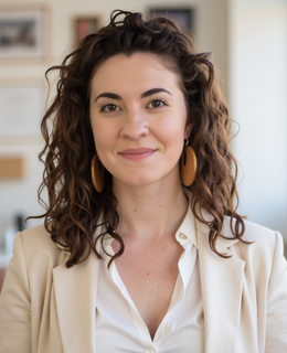 Christina has brown curly hair, brown eyes, and is wearing a white shirt and tan blazer.