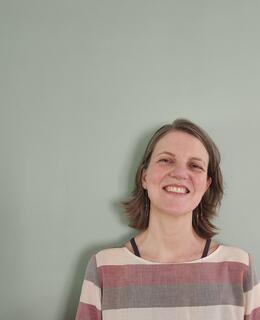 the image shows the upper body and head of a middle aged, scandianavian woman leaning up against a dustry blue wall. She is smiling and her neck length dark blond hair hangs freely. She is wearing a loose, striped shirt in grey and rose tones.