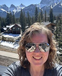 A woman wearing sunglasses smiles standing in front of the Three Sisters Mountain