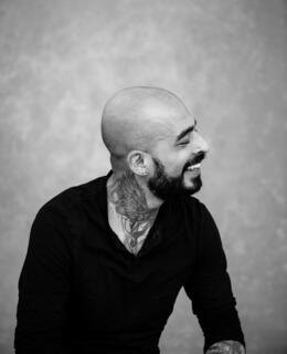 Grayscale image of a bald, South Asian man with a beard looking to the right while smiling. He is wearing a blackshirt and has a throat tattoo - it is a pair of wings in the Trash Polka style.