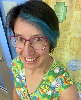 Woman with short black hair with bright blue highlights wearing bright clothing and glasses smiling at the camera.