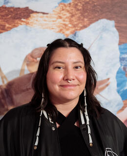 Person in all black wearing dentalium shell earrings. Standing in front of a blue, white, and copper painting. 