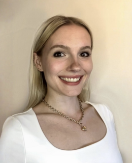 Aimee is wearing a white t-shirt and gold heart necklace. She is a white woman with blonde, straight hair and brown eyes.