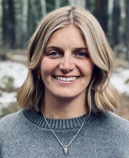 Blonde woman wearing a grey sweater standing in front of a forest background