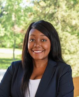 A headshot of a Black women smiling and standing behind a scenic background