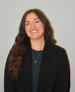 A photo of a woman with dark brown hair. She is wearing a green shirt with a black blazer on top. 