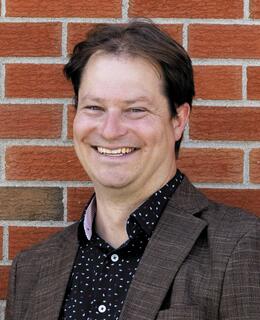 White male face smiling in front of bring wall. Brown blazer with blue button up shirt.